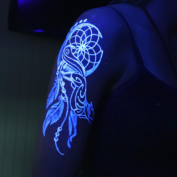 Glow in the dark tattoo of intricate feathers and geometric design, visible under black light on a person's arm.
