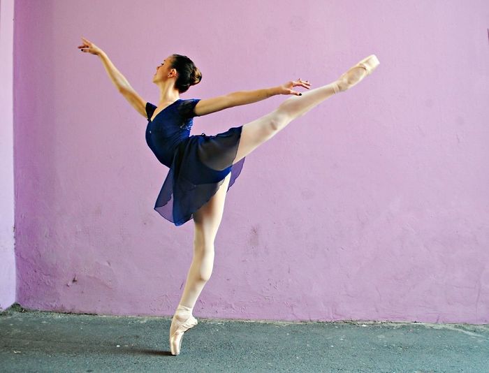 Ballerina in a blue dress performing an arabesque against a purple wall for Ballet Day.