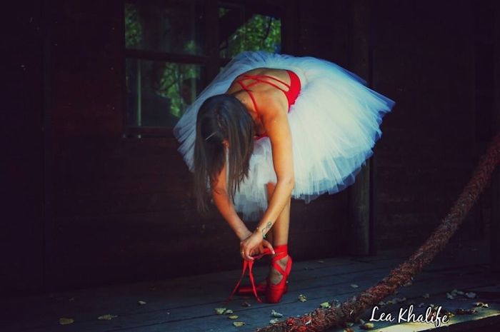 Ballet dancer in a white tutu adjusting red pointe shoes in a dimly lit studio.