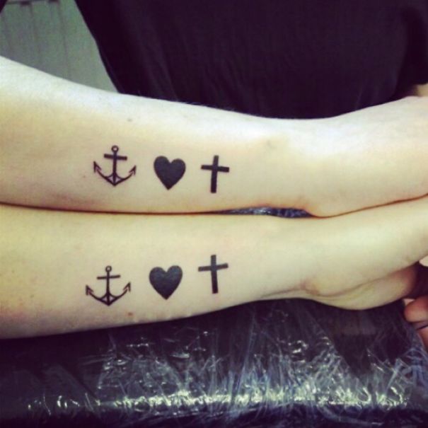 Two arms with matching sister tattoos featuring an anchor, heart, and cross design, symbolizing a strong bond.