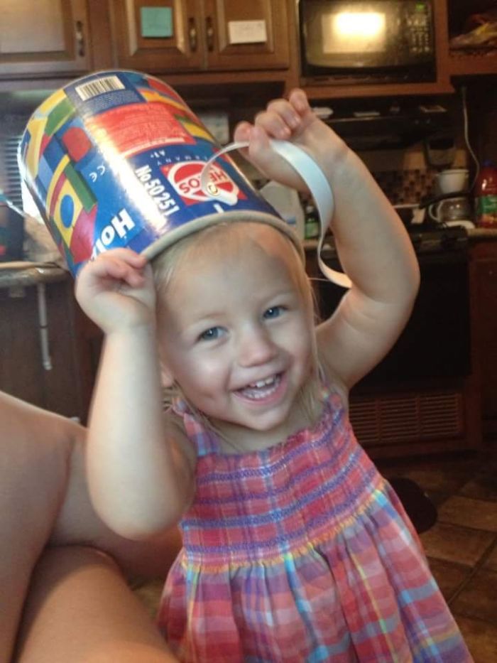 Toddler wearing a colorful bucket on her head, smiling and playing indoors, showing kids as copy-paste versions of their parents.