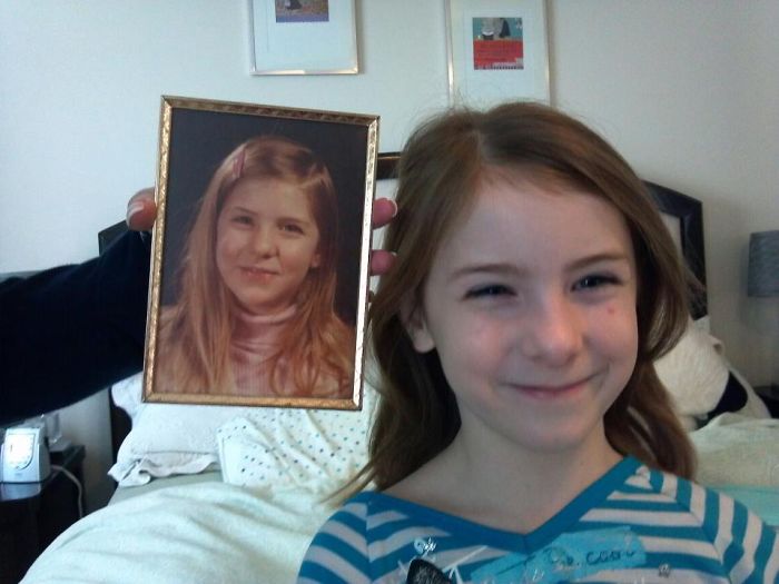 Young girl smiling next to a framed photo of her mother, showing striking copy-paste resemblance between kids and parents.