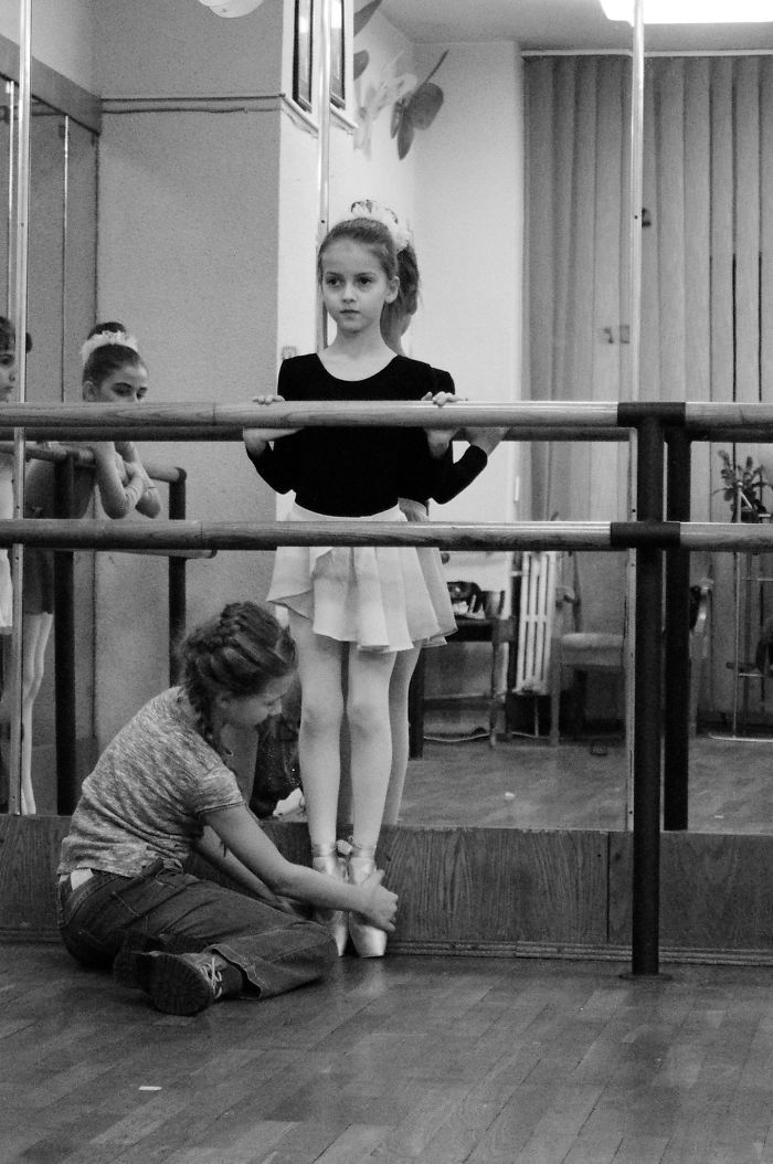 Young ballerina at the barre, teacher adjusting her pointe shoes, celebrating Ballet Day.