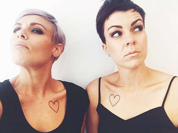 Two women with matching heart sister tattoos on their chests, wearing black tops.