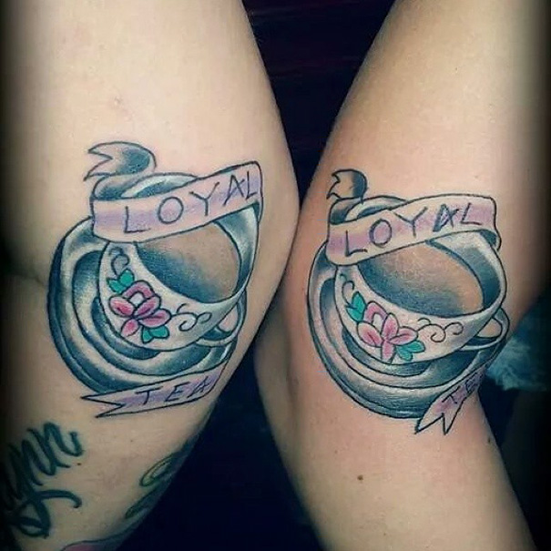 Matching sister tattoos of teacups with "loyal" ribbon and floral design.