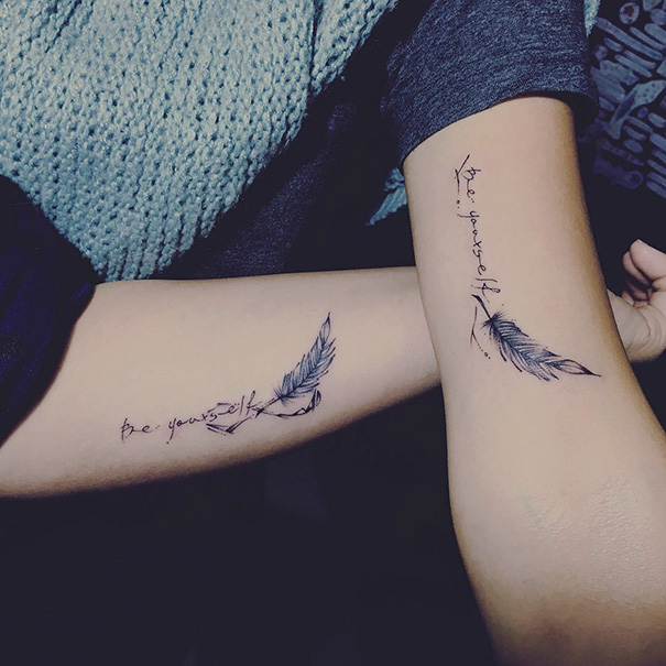 Matching sister tattoos on arms with feathers and "be yourself" text.