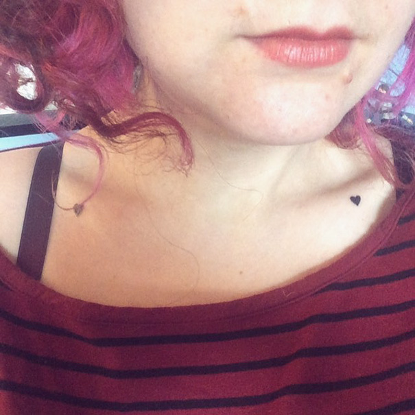 Minimalist tattoo of a small heart near the collarbone on a person with curly pink hair, wearing a striped top.