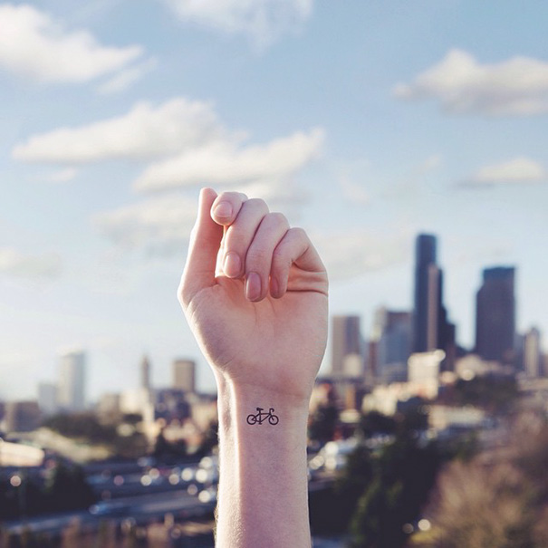 Hand with a minimalist bicycle tattoo in front of a city skyline, showcasing minimalist tattoos.