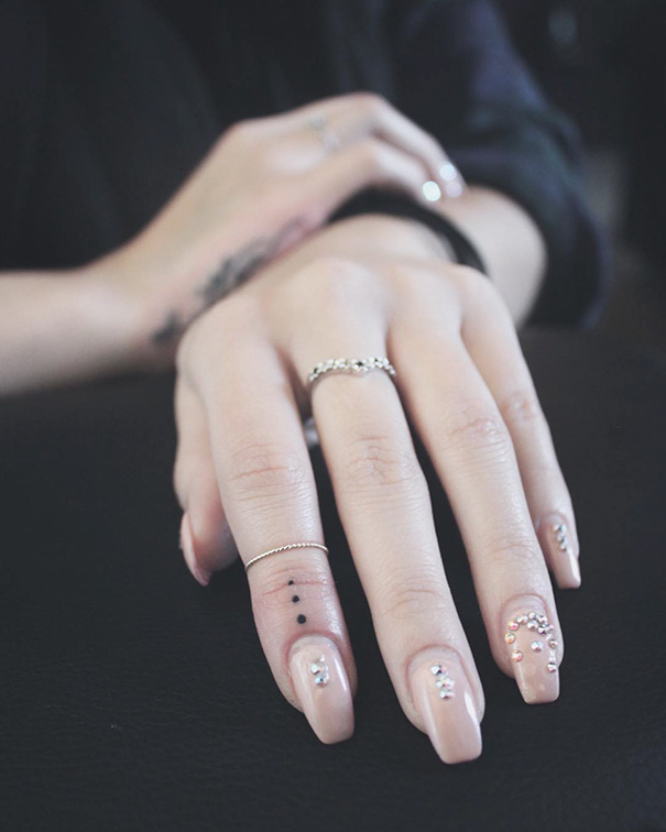Hand with minimalist dot tattoo on ring finger, wearing delicate rings.