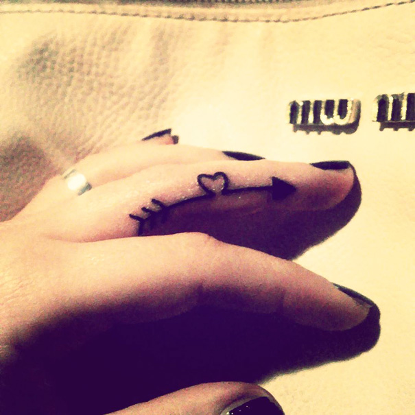 Minimalist tattoo of arrow with heart on a finger, black nail polish visible.