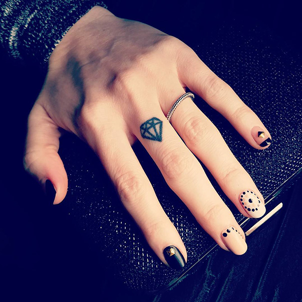 Minimalist tattoo of a diamond on a finger, hand resting on a textured surface with black and white nail art.