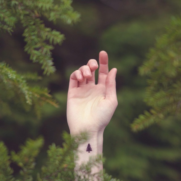 Minimalist tattoo of a small tree on a wrist, surrounded by green foliage.