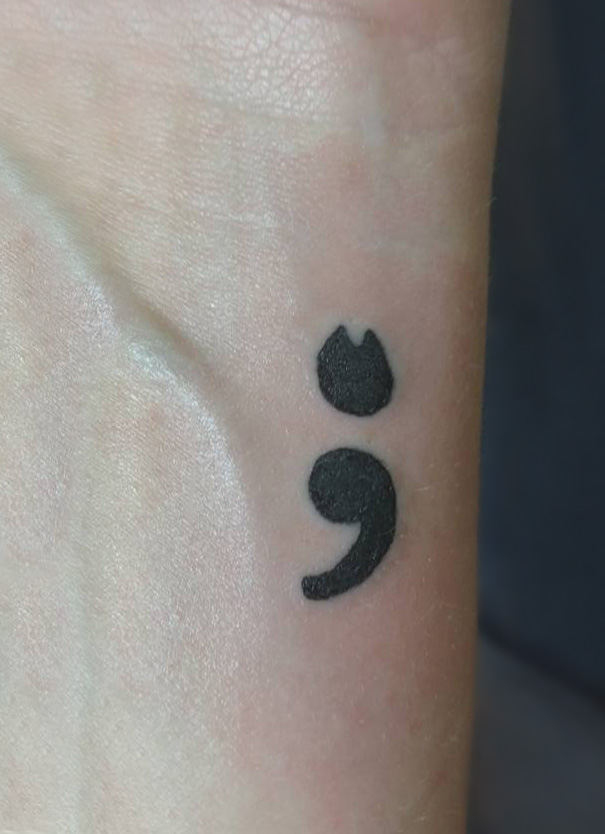 Minimalist tattoo of a semicolon and cat paw on a wrist.