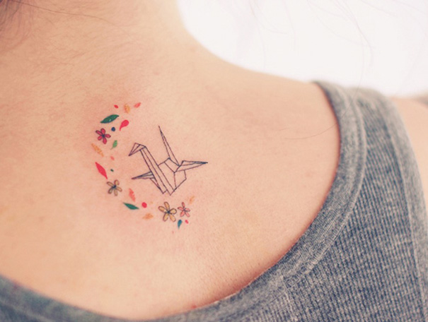 Minimalist tattoo of an origami crane with colorful floral accents on the back of a neck.