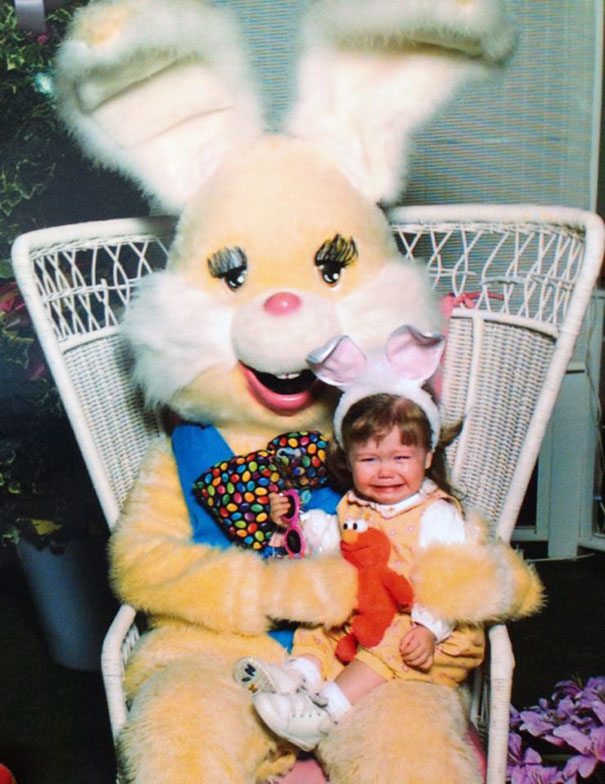A child sitting nervously on a giant vintage Easter bunny's lap, both in a wicker chair.