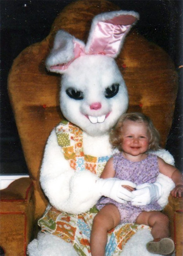 Smiling child sitting with a vintage Easter bunny in a colorful outfit on a brown chair.