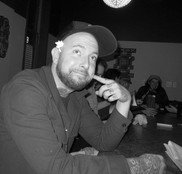 Tattoo artist offers free cover-up tattoos, seated with others in a casual setting, wearing a cap and jacket. Tattoo artist offers free cover-up tattoos, seated with others in a casual setting, wearing a cap and jacket.