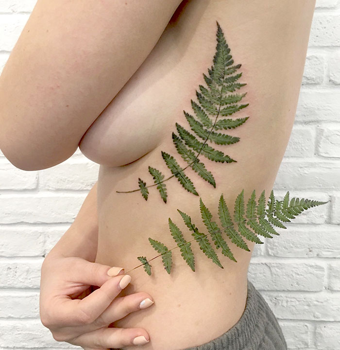 Person with a botanical tattoo of a fern leaf on their side, using a real leaf as a stencil.
