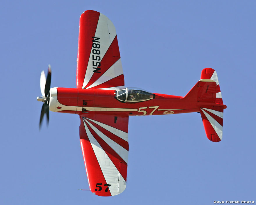 Red and white aircraft with propeller, flying against a clear sky, showcasing dynamic aerial design.