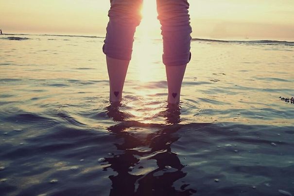 Person standing in shallow water at sunset, showing small foot tattoos with heart designs.