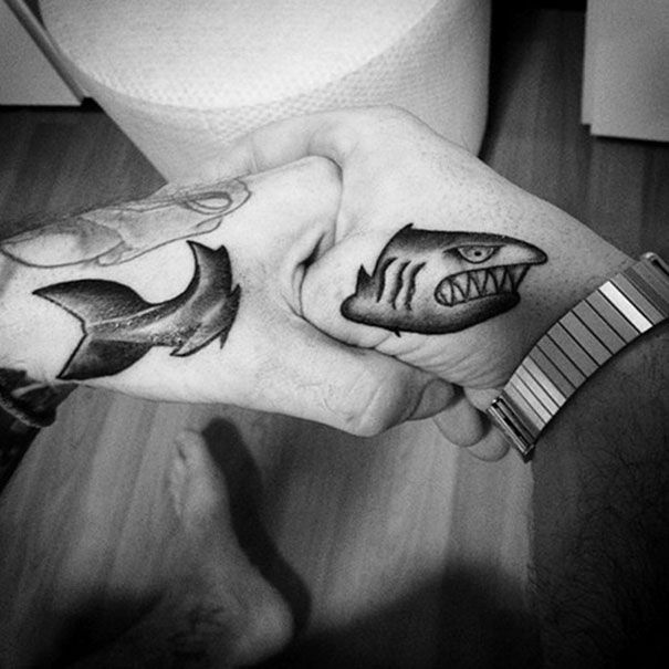 Two hands with clever tattoos of shark halves creating a complete shark when fists are placed together.