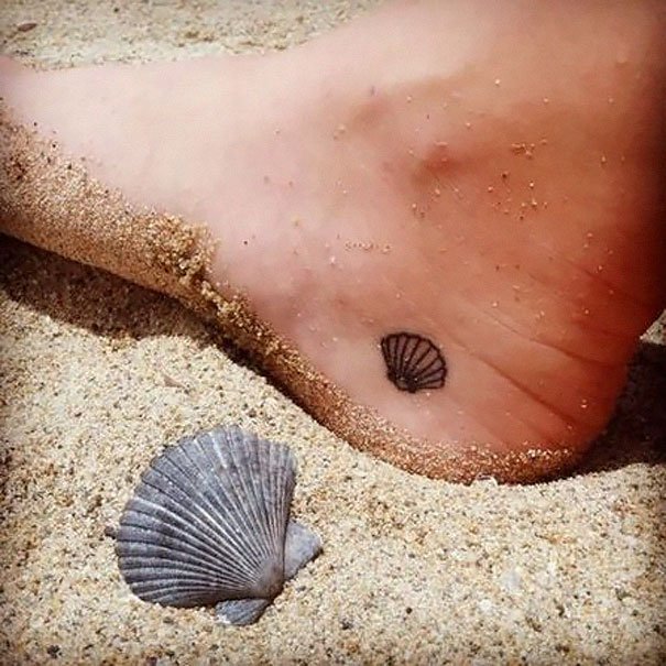 Small foot tattoo of a shell, partially covered by sand, next to a real shell on a beach.