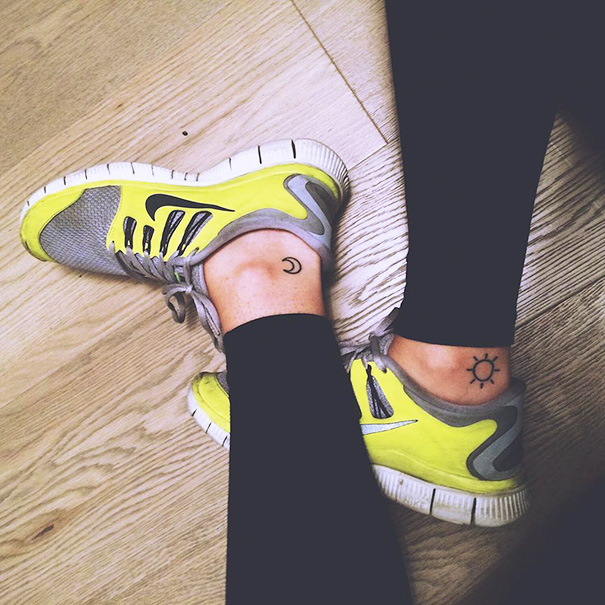 Small foot tattoos of a moon and sun on ankles, wearing lime green sneakers on wooden floor.