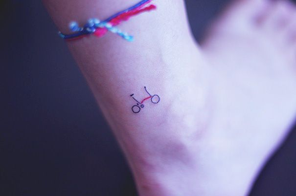 Small foot tattoo of a minimalist bicycle with a colorful ankle bracelet.