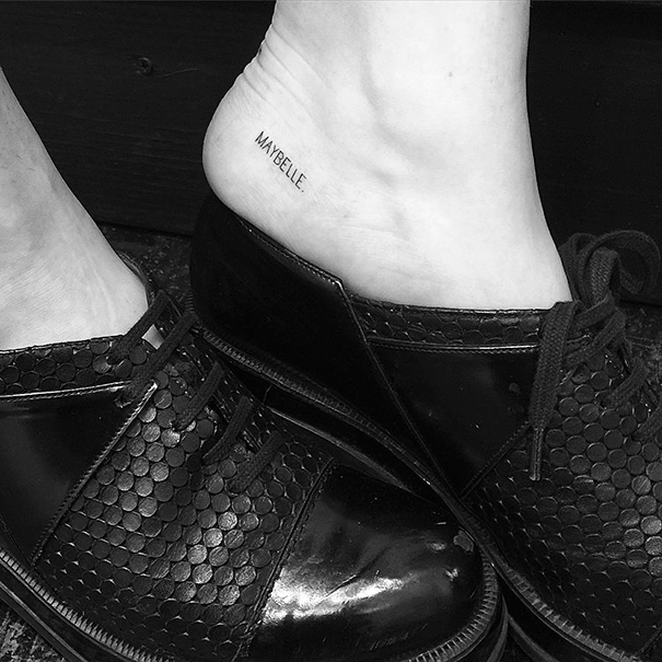 Small foot tattoo reading "Maybelle" above black shoes, showcasing minimalist tattoo design.