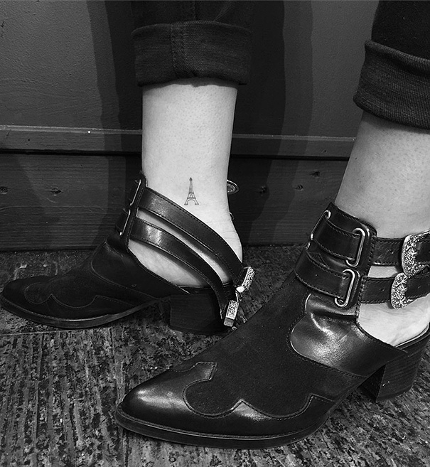 Minimalist small foot tattoo of Eiffel Tower on ankle, showcasing a "less is more" design approach in black and white.
