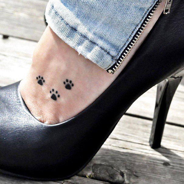 Small foot tattoo of tiny paw prints, a minimalist design on a woman's foot in high heels and denim.