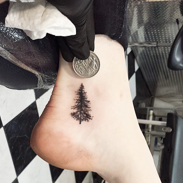 Small foot tattoo of a tree, showcasing minimalist design with a coin for size comparison on a checkered floor.