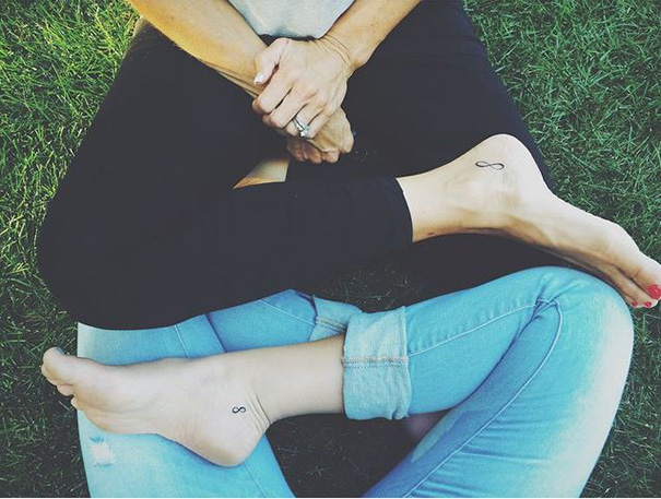 Two feet with minimalist small foot tattoos of infinity symbols resting on grass.