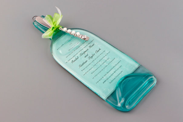 Creative wedding invitation made from a flattened recycled glass bottle with printed text and decorative beads on a gray background.