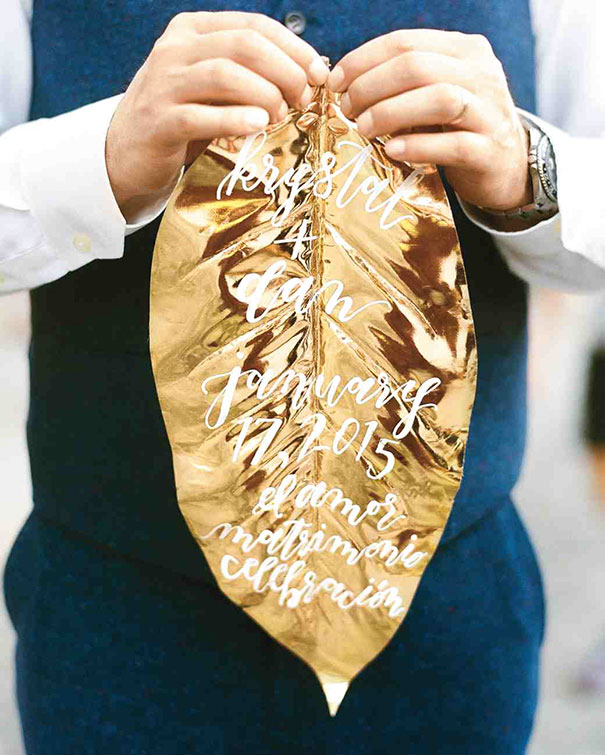 Person holding a shiny gold leaf creative wedding invitation with white calligraphy text.