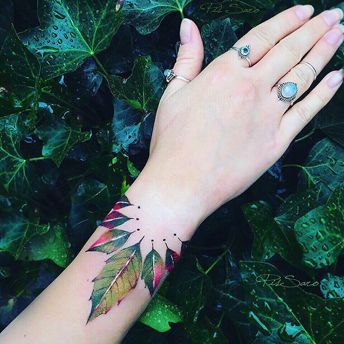 Ethereal nature tattoo on wrist with leaves in vibrant seasonal colors, surrounded by green foliage.
