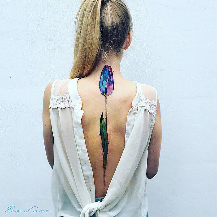 Back view of a woman with an ethereal nature tattoo of a colorful flower, inspired by changing seasons, on her spine.