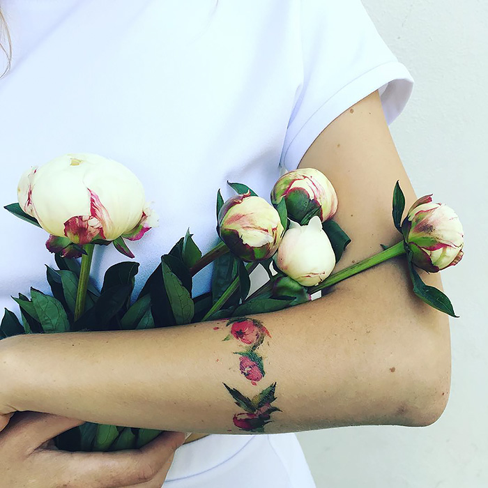 Person holding peony flowers with a floral tattoo on their arm, showcasing ethereal nature-inspired design.