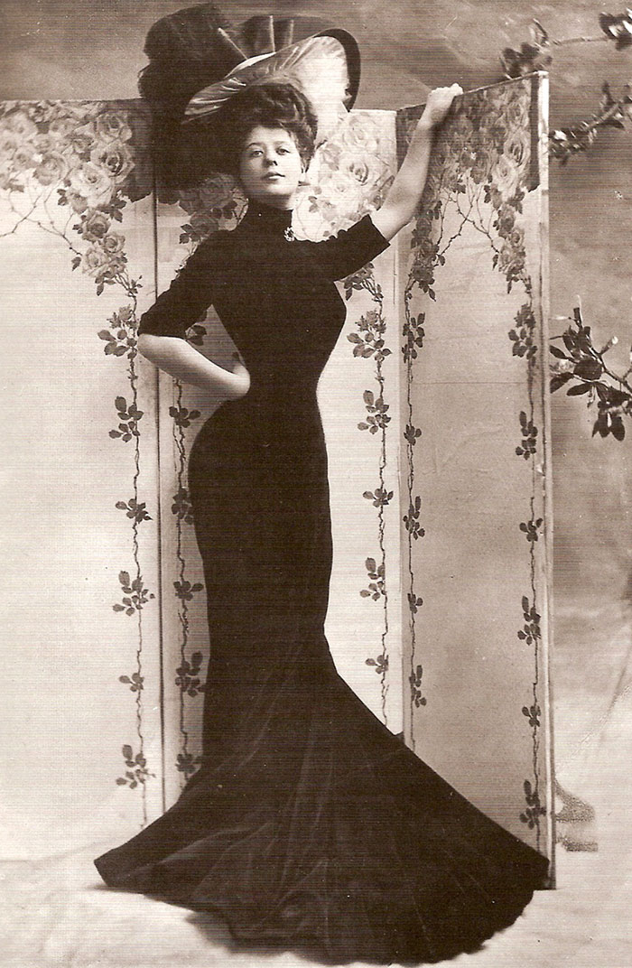 Edwardian era woman in elegant dress and large hat, posing with floral backdrop.