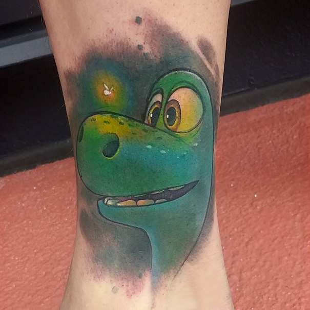 Pixar-inspired tattoo of a smiling green dinosaur with a glowing firefly on a forearm.