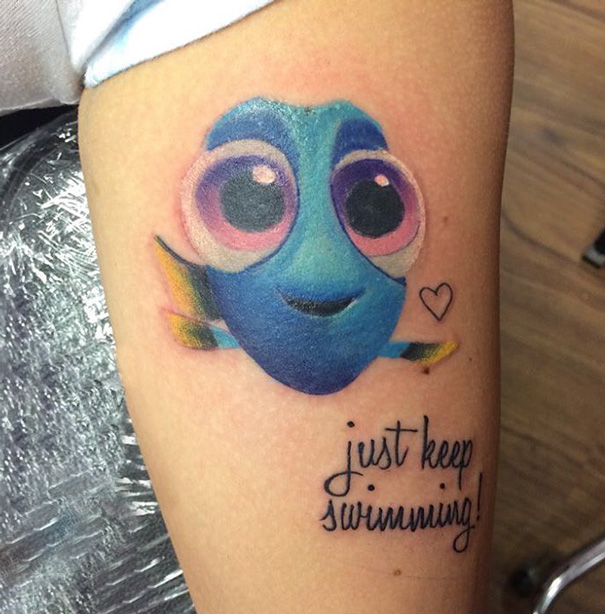 Pixar-inspired tattoo of a blue fish with large eyes and the phrase "just keep swimming" on an arm.