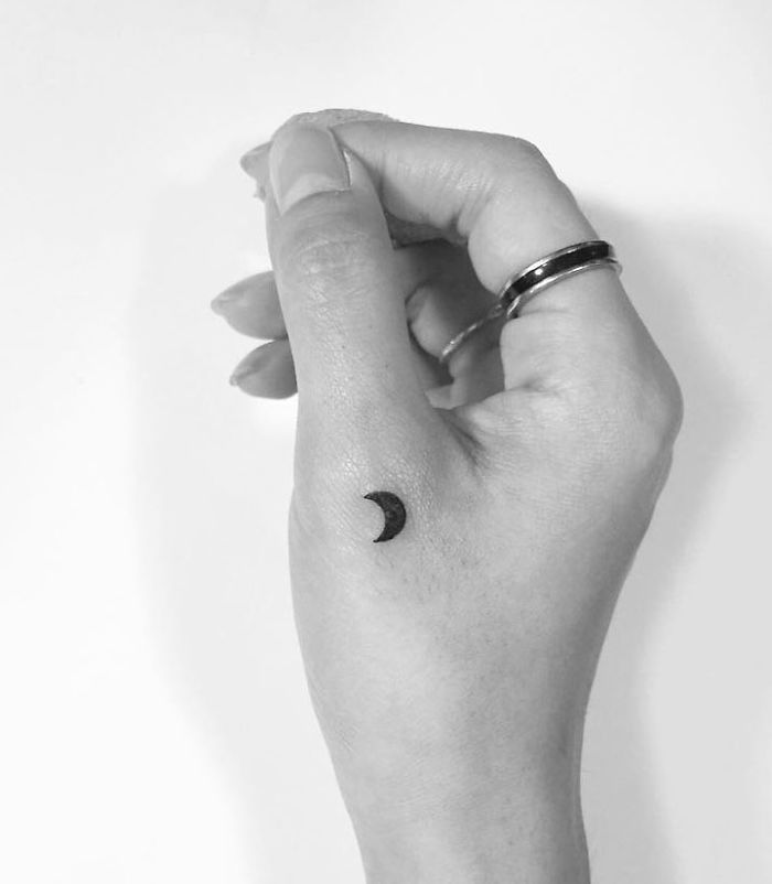 Minimalist tattoo of a small crescent moon on a person's hand, designed by a Korean artist.