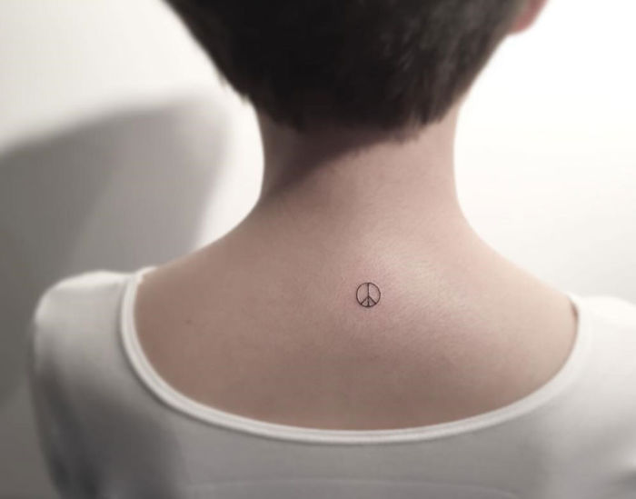Minimalist tattoo of a peace symbol on the back of a person wearing a white shirt, created by a Korean artist.