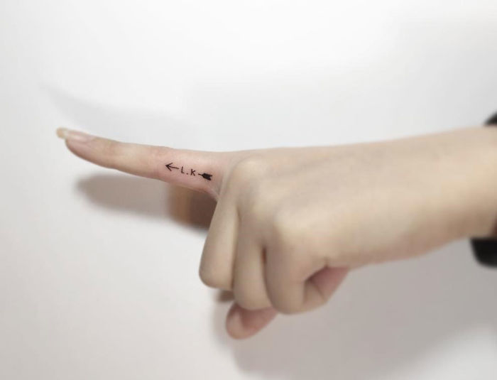 Minimalist tattoo by Korean artist on finger, featuring small arrow design with initials.