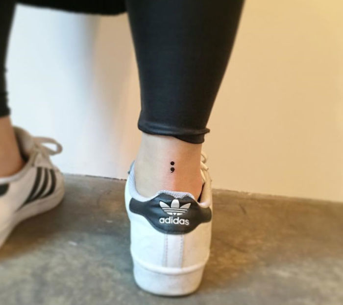 Minimalist tattoo of a semicolon on the ankle, near Adidas shoe, designed by a Korean artist.