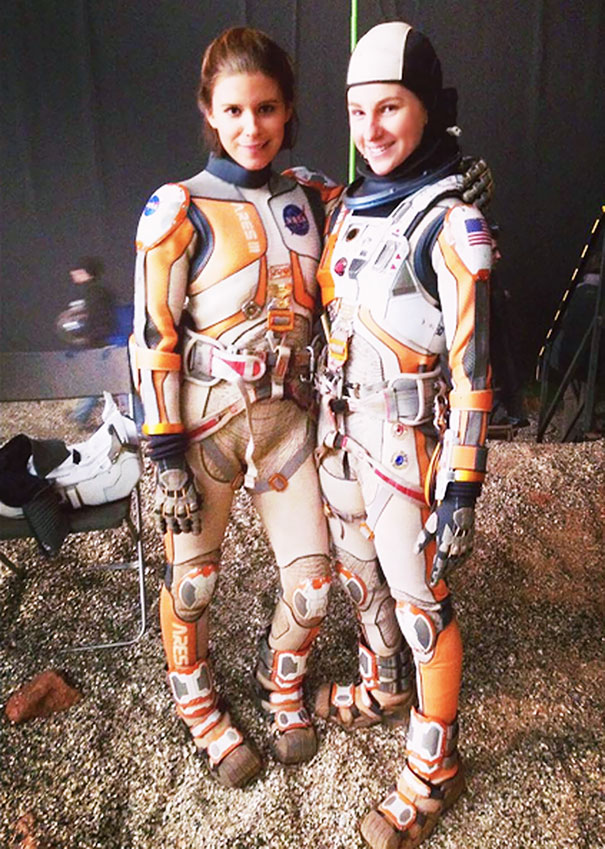 Two women in detailed orange and white space suits posing together as celebrity body doubles on a Hollywood set.