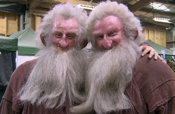 Two celebrities’ body doubles dressed as elderly men with long white beards posing together on a film set.