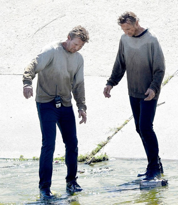 Two men dressed alike in dirty sweatshirts and dark pants standing in shallow water, showcasing celebrity body doubles.