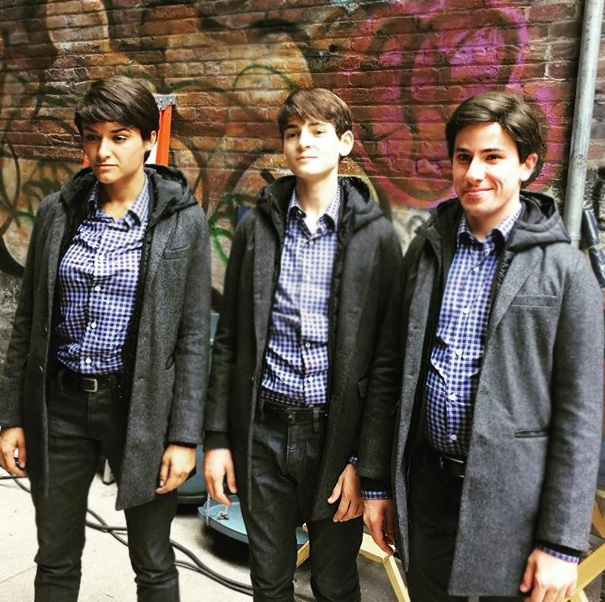 Three people wearing matching checkered shirts and gray coats standing in front of a graffiti wall illustrating celebrity body doubles.