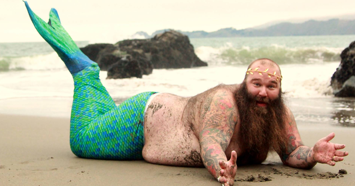 Bearded man with tattoos wearing a green and blue mermaid tail posing for a dudeoir photoshoot on the beach.